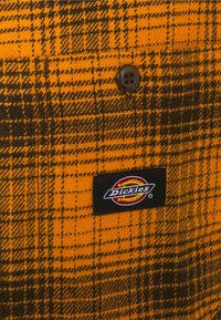 Bouton noir sur tissu à carreaux orange et noir avec une étiquette de marque Dickies cousue près du bas au centre du tissu.