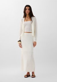 Witte gebreide cropped tanktop onder een kanten cardigan, gecombineerd met een lange witte rok met textuurelementen. Bruine sandalen met gouden accenten.