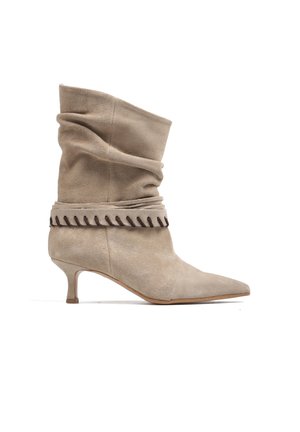 Beżowy zamszowy botek do połowy łydki z luźną cholewką, szpiczastym noskiem, niskim obcasem typu kitten heel oraz ozdobnym przeszywanym paskiem wokół kostki.