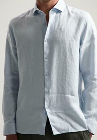 Camisa de botones de manga larga color azul claro en tela de lino, con cuello puntiagudo, tapeta frontal con botones y pequeño logo en el pecho.