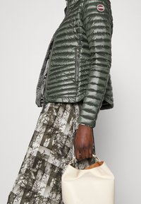 Tmavě zelená prošívaná bunda s vodorovným vzorem, se zipovými kapsami, nošená přes vzorovanou sukni. Ruka drží krémovou kabelku.