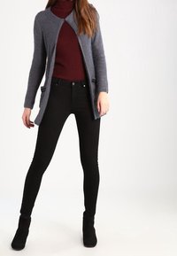 Žena v šedém otevřeném kardiganu, burgundské roláku, černých slim kalhotách a černých kotníkových botách, stojící s nohama od sebe.