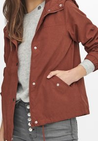 Rust-colored jacket with a zip front, hood, and snap buttons. Features kangaroo pockets and ribbed gray cuffs, paired with a gray sweater.