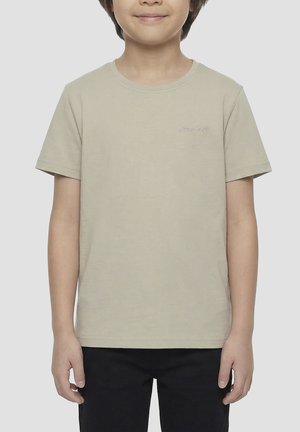 Kind draagt een effen beige T-shirt met korte mouwen en een zwarte broek, staand met ontspannen armen tegen een neutrale achtergrond.