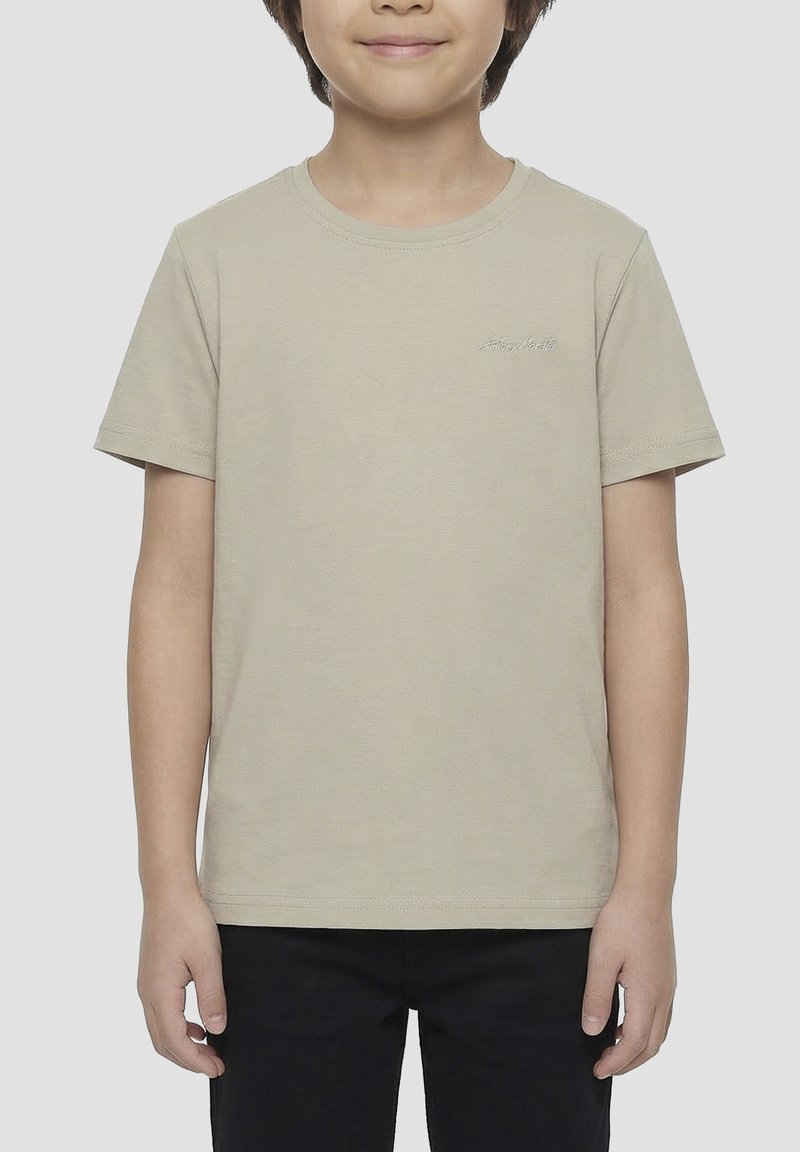 Enfant portant un t-shirt beige uni à manches courtes et un pantalon noir, debout les bras détendus devant un fond neutre.