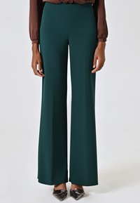Pantalons larges verts en tissu lisse, dotés d'une taille haute et d'une coupe confortable, associés à des chaussures noires à talons pointus.