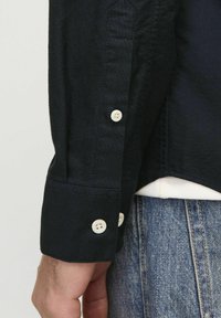 Marineblaues Hemd mit langen Ärmeln und weißen Knöpfen; verfügt über einen strukturierten Stoff und ein schlankes Manschettendetail in der Nähe des Handgelenks.