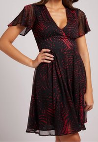Robe noire avec motif de feuilles rouges ; décolleté en V, manches courtes en volants, corsage ajusté et jupe plissée. Fabriquée en tissu léger et semi-transparent.