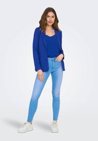 Blazer azul sobre una blusa azul, combinado con jeans skinny de denim claros. Zapatillas blancas completan el atuendo. Tela suave, diseño a medida.