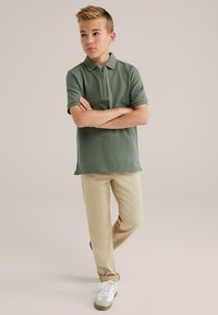 Zelené polo tričko s krátkým rukávem a límcem na zip, ve spojení s béžovými kalhotami a bílými teniskami, nastavené na neutrálním pozadí.