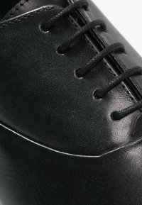 Gros plan sur une chaussure de ville en cuir noir poli avec des lacets noirs passés à travers les œillets, mettant en valeur la texture et les coutures.