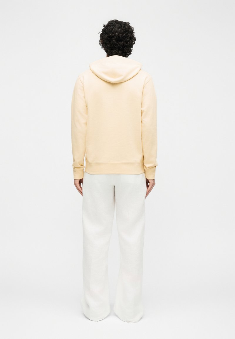 Person with curly hair stands facing away, wearing a light beige hoodie and loose white pants against a plain background.