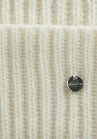 Cremfarbener, gerippter Strickstoff mit einem runden silbernen Anhänger, auf dem der Text "capsule" steht, und der ein strukturiertes und mehrschichtiges Design hervorhebt.