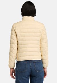 Lys gul pufferjakke, quiltet design med vandrette sektioner, høj krave og tætsiddende ærmer, parret med blå jeans.