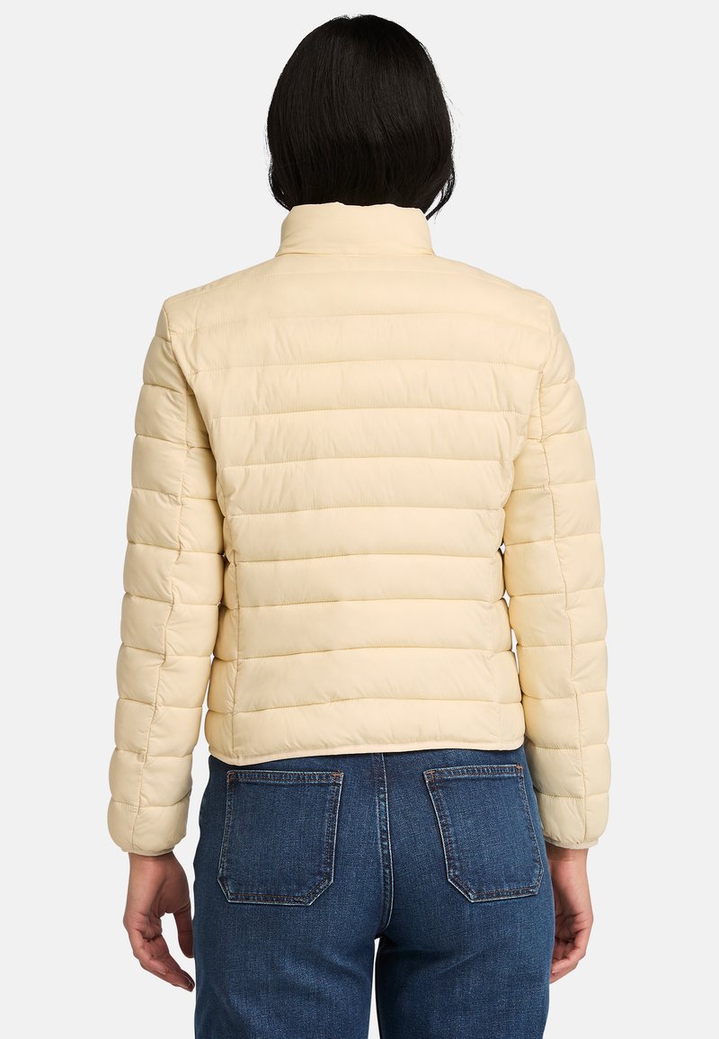 Giacca puffer gialla chiara, design trapuntato con sezioni orizzontali, colletto alto e maniche aderenti, abbinata a jeans blu.