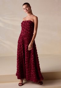 Bordeauxrode strapless jurk met een gestructureerd rozenpatroon. Vloeiende, tot op de vloer reikende rok. Simpele sandalen maken het ontwerp af.
