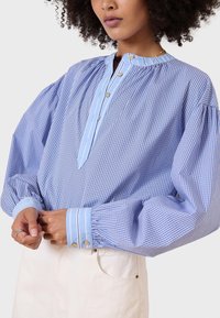 Camicia a quadri blu e bianca con colletto alto, bottoni sulla placca e maniche lunghe a palloncino. Accentuata con polsini e righe azzurre.