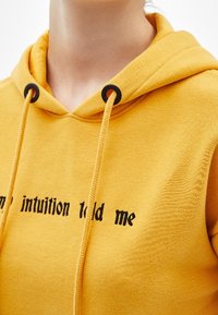 Primer plano de una sudadera amarilla con ojales de cordón negros y un texto negro parcial que dice "mi intuición me lo dijo" en el pecho.