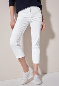 Jeans blancos recortados de mezclilla, con un ajuste slim, bolsillos frontales y cierre de botón. Combinados con zapatillas blancas con cordones.