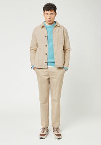 Beige canvas jacket with two front pockets, paired with a turquoise knit sweater and light beige trousers, completed with brown and white sneakers.