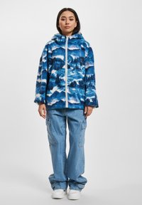 Fleece hette med blå og hvit landskapsmønster, glidelås foran og hette; kombinert med lyseblå cargojeans og hvite joggesko.