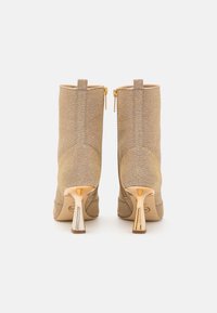 Botines de tobillo en tono dorado, hechos de tejido texturizado, con cierre de cremallera y un distintivo tacón dorado con un diseño geométrico.