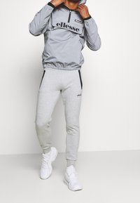 Šedá větrovka s polo-zip designem a černým logem, v kombinaci se šedými tepláky se bočními kapsami a brandováním. Bílé tenisky.