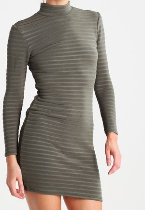 Eng anliegendes olivgrünes, langärmeliges, geripptes Kleid mit hohem Kragen, das bis zur Mitte des Oberschenkels reicht, getragen von einer Person vor einem einfarbigen Hintergrund.