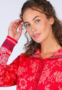 Roter Pullover mit Blumenmuster und Reißverschlusskragen, ausgestattet mit kontrastierenden gemusterten Bündchen in Marineblau und Weiß. Weicher Stoff mit Knopfdetails.