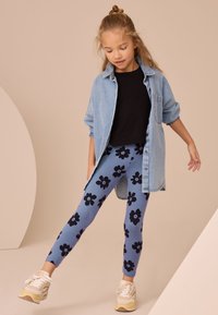 Kék leggings fekete virágmintával, farmeringgel és fekete felsővel párosítva. A cipők krémszínűek és bézs árnyalatúak, fűzővel.