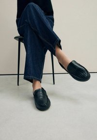 Zwarte leren instappers met een textuurpatroon van krokodillenleer, met een ronde neus en gestikte randen, gedragen met donkere jeans.