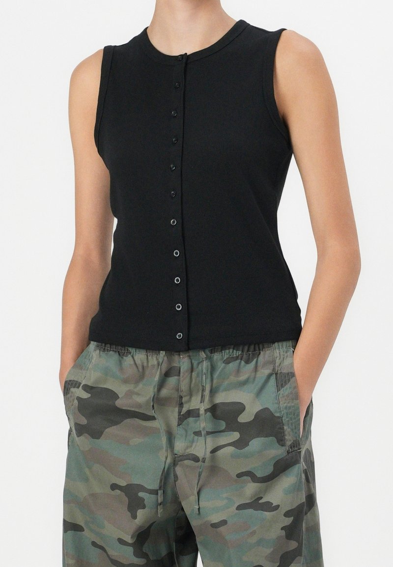 Haut noir sans manches avec un col rond et des boutons sur le devant ; en tissu côtelé. Associé à un pantalon cargo à motif camouflage clair.