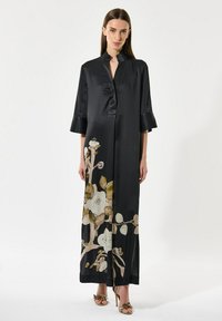 Vrouw gekleed in een lange zwarte satijnen jurk met brede mouwen en een beige bloemenprint vlakbij de zoom, gecombineerd met gouden hoge hakken.