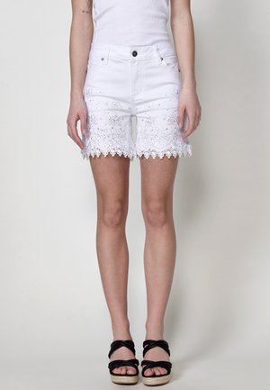 Shorts en denim blanc avec des détails en dentelle florale au bas, un bord festonné et cinq poches avec une fermeture à bouton noir.