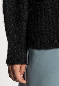 Maglione nero lavorato a maglia con una finitura testurizzata e pelosa; presenta maniche lunghe e un orlo a costine, indossato sopra un indumento azzurro chiaro.