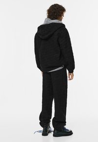 Giacca a vento nera imbottita con cappuccio e fodera a quadri, abbinata a pantaloni imbottiti coordinati e stivali neri con lacci. Dettaglio del tessuto testurizzato visibile.