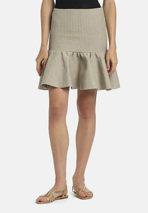 Beiger, gerippter Strickrock mit ausgestelltem Saum, kombiniert mit goldfarbenen, flachen Riemchensandalen, getragen von einem Modell vor neutralem Hintergrund.