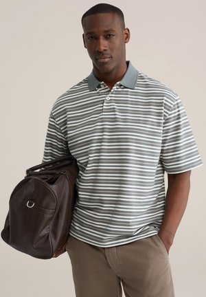 Muž v sivej a bielo pruhovanej polokošeli a béžových nohaviciach, drží na ramene veľkú tmavohnedú koženú tašku cez plece.