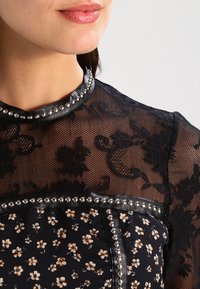 Schwarzes Spitzentop mit hohem Kragen, floralen Mustern und dekorativen silbernen Nieten entlang des Kragens und des Oberteils für zusätzliche Details.