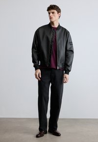 Juoda bombonešio striukė su lygiu paviršiumi, ribuotu rankogaliu ir apykakle. Dėvima ant bordo spalvos marškinio, derinama su tamsiomis džinsais ir rudos spalvos batais.