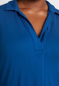 Close-up of a person wearing a blue collared shirt with an open neckline showing upper chest skin.