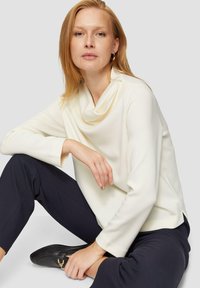 Haut à col châle, à manches longues en tissu crème. Texture douce, coupe décontractée, fentes latérales ; associé à un pantalon bleu marine foncé et des chaussures slip-on noires.