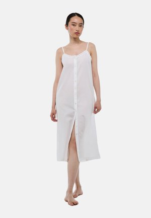Robe blanche sans manches avec un devant à boutons, bordée de dentelle au niveau du décolleté, et une fente haute sur la cuisse pour un mouvement facilité. Texture de tissu lisse.