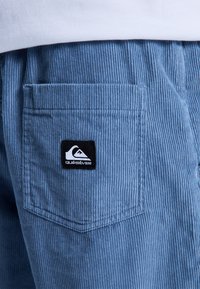 Pantalons en velours côtelé bleus avec une poche arrière ornée du logo noir Quiksilver. La texture est cannelée, ajoutant de la profondeur au design du tissu.
