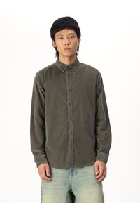 Chemise boutonnée en velours côtelé vert olive avec un col évasé, une poche poitrine et des boutons foncés, associée à un jean en denim bleu clair.