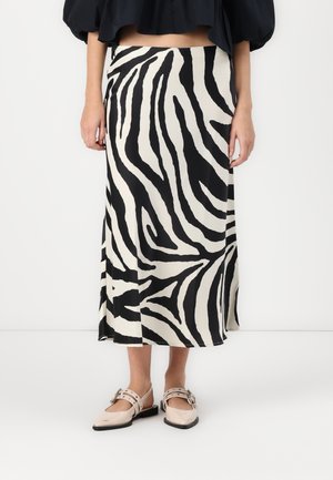 Vrouw draagt een midi rok met zebraprint, een zwart cropped top en beige Mary Jane schoenen, staand tegen een effen witte achtergrond.