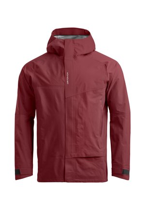 Mann mit grüner Mütze, kastanienbrauner Jacke, schwarzer Hose und grün-schwarzen Wanderschuhen, der den Reißverschluss an der Vorderseite der Jacke schließt.