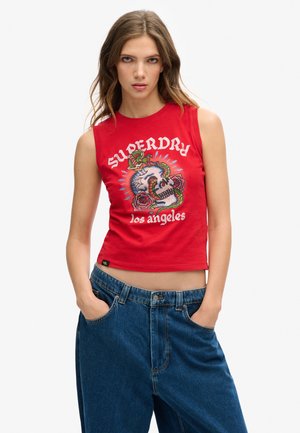 Débardeur rouge sans manches avec un imprimé graphique représentant un crâne, des roses et le texte "SUPERDRY los angeles". Porté avec un pantalon en denim bleu ample.