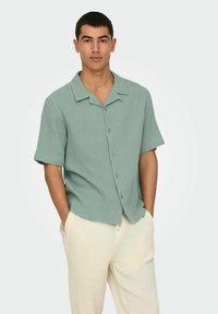 Chemise verte clair à manches courtes avec un tissu texturé, fermeture à boutons et une coupe décontractée, associée à un pantalon ample de couleur crème.