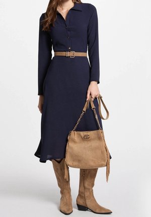 Marineblå langærmet kjole med krave og knapper foran, kombineret med et tanfarvet bælte og håndtaske. Modellen bærer knæhøje tanfarvede støvler.